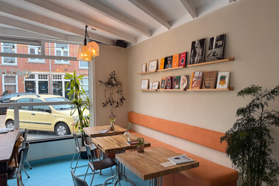 The interior of a bright, minimalist cafe with white wooden ceiling beams and light blue painted floorboards. The shot focuses on the service counter with a large coffee grinder and espresso machine. To the right, customers sit at a high table near a large window, and large green plants are placed around the room.