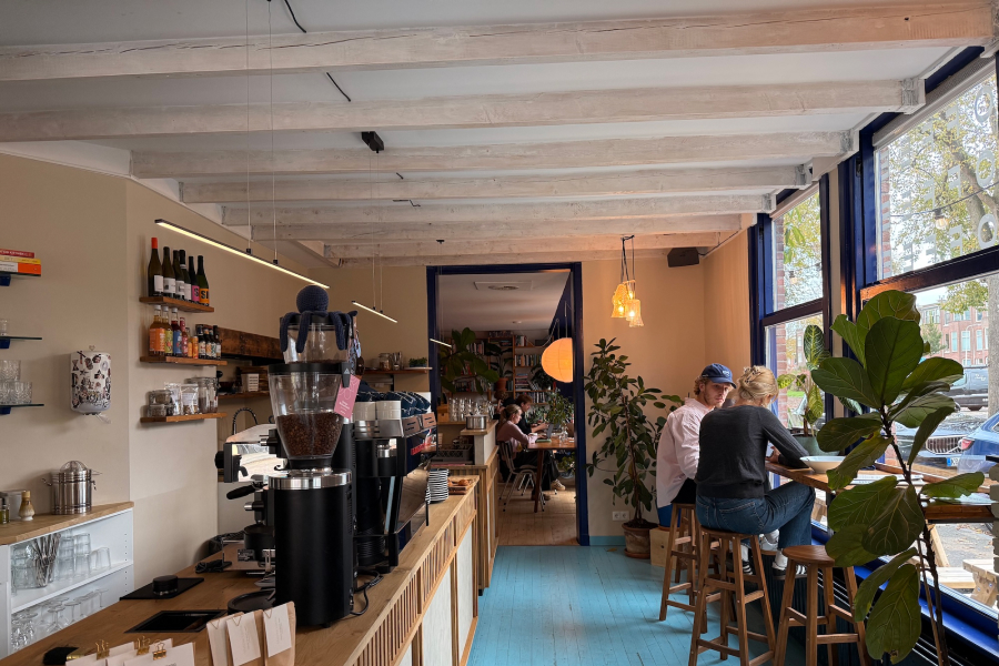 A view of the seating area in a bright cafe. The room has light blue floorboards, small wooden tables with dark metal chairs, and a built-in bench with orange upholstery along the wall. Above the bench, a wooden shelf displays a collection of books and small framed artwork. A large window shows a street scene with parked cars.