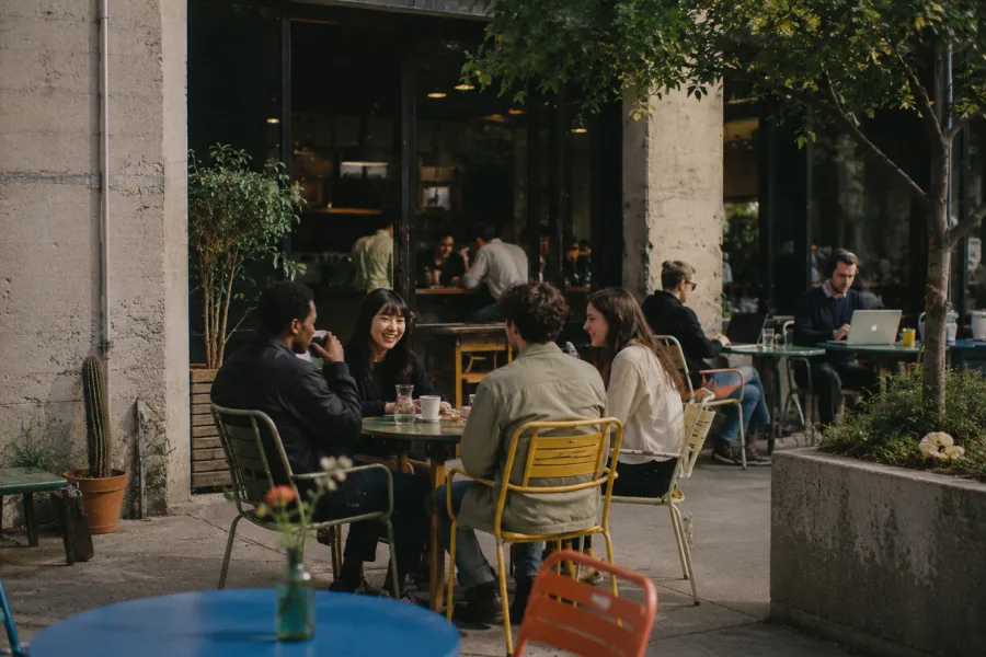 A diverse group of friends sit at a colorful outdoor cafe table, drinking coffee and chatting in front of a modern concrete cafe. Around them, other visitors work on laptops, with greenery and bright furniture adding a relaxed, urban atmosphere.